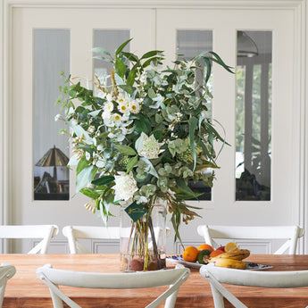 A large artificial floral arrangement featuring a variety of green and white flowers in a dome-shaped vase, placed on a table with chairs around it.